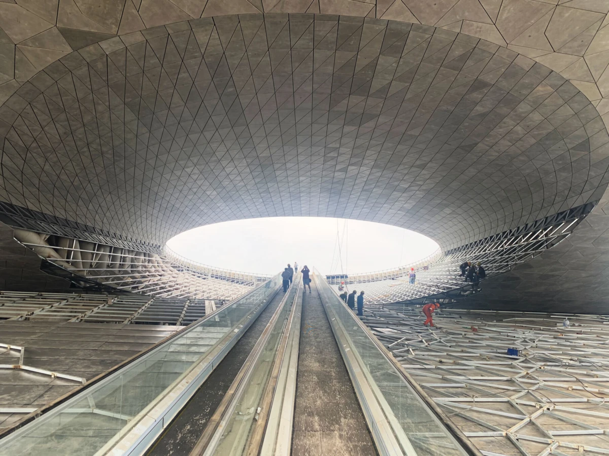 Escalator ascending toward a bright circular opening in a domed, geometric concrete atrium; several people on the walkway.