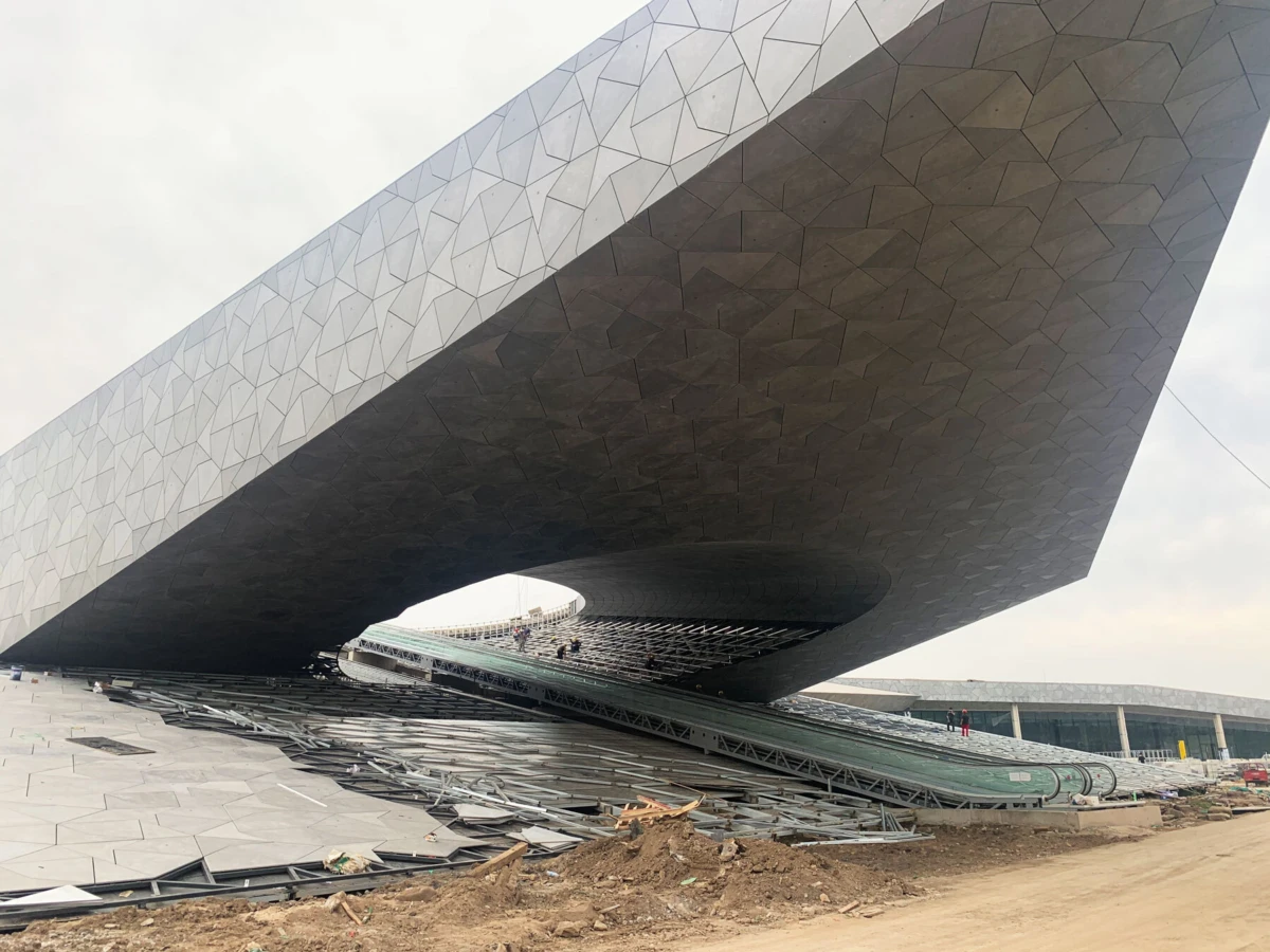 Massive cantilevered structure with tessellated metal panels; exposed underside and elevated walkway amid construction debris.