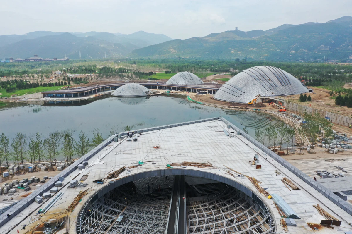 Under-construction complex of dome-shaped pavilions along a lakeside, with a large circular framework in the foreground and distant mountains.