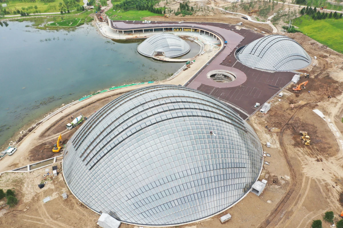 A cluster of glass-domed geodesic structures under construction along a lakeshore, connected by brown-paved walkways.