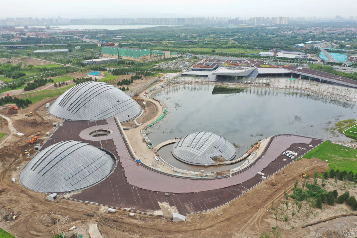 Aerial view of a glass-domed architectural complex under construction, featuring three interconnected domes and a curved walkway circling a pond.