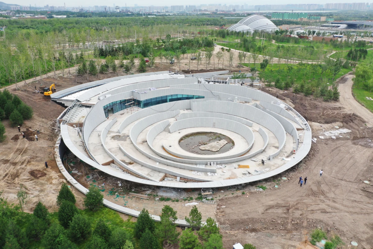 Concentric ring-shaped concrete structure under construction in a green park, with excavator and workers nearby, distant city skyline.