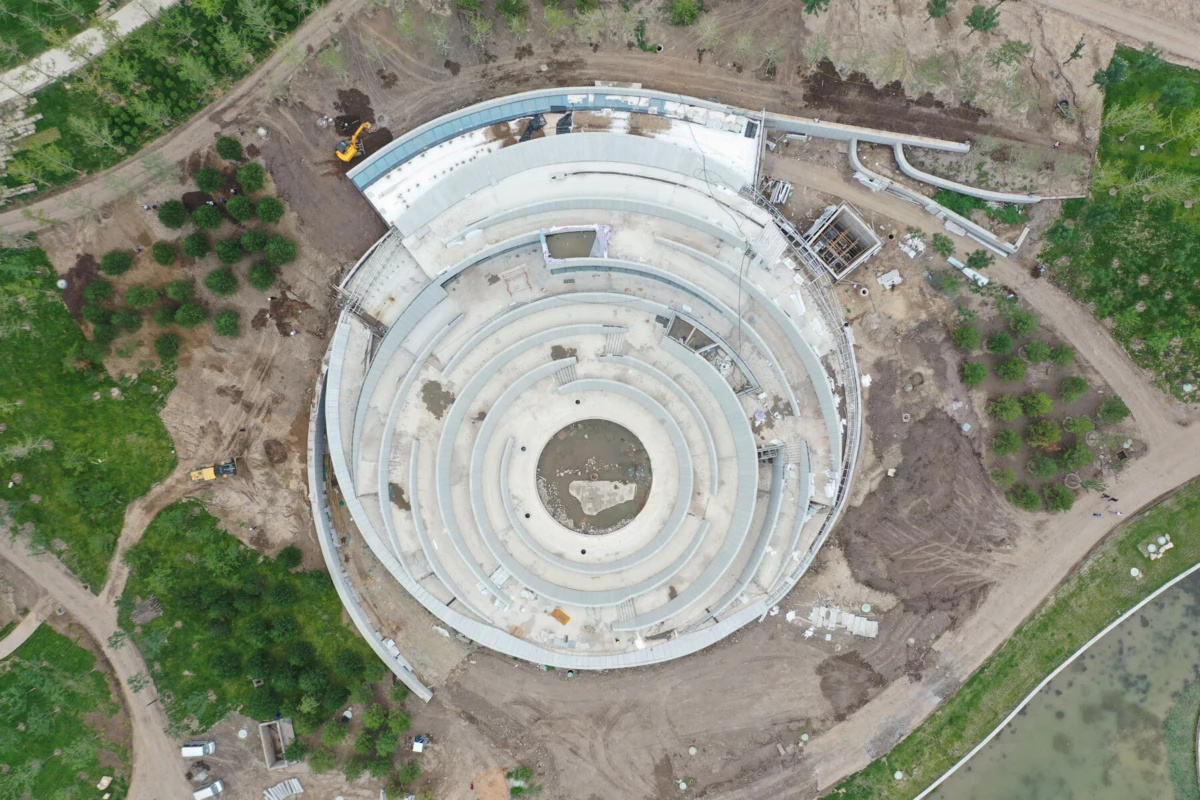 Aerial view of a circular spiraling concrete structure under construction, surrounded by trees and construction equipment.