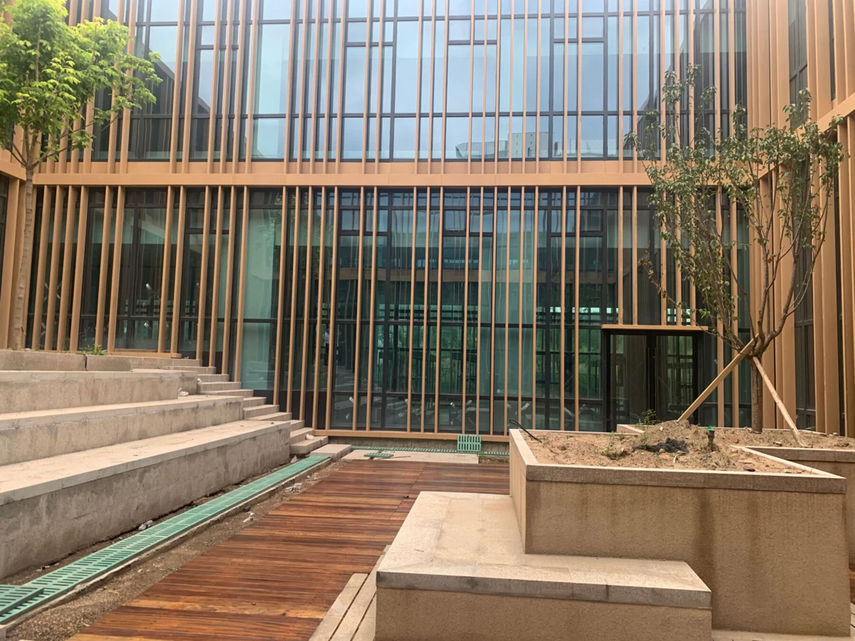 Empty courtyard in front of a glass-fronted building with vertical wooden louvers, concrete steps, and a raised planter with a tree.