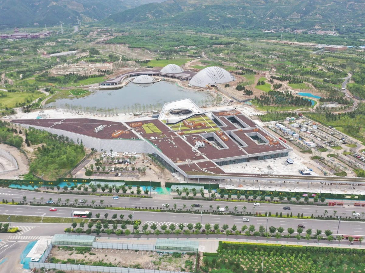Aerial view of a large modern complex with a star-shaped building around central courtyard, dome-roof pavilions by lake, and highway in foreground.