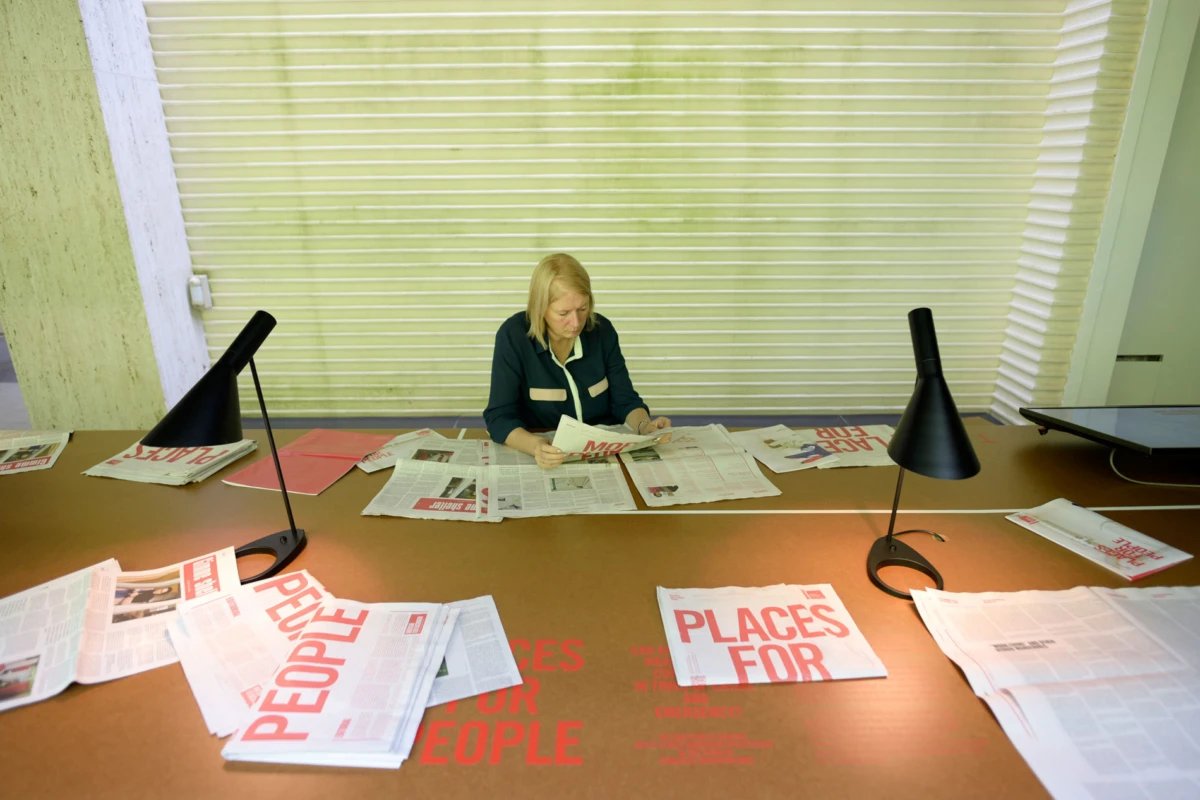 Blonde-haired person sits at a long desk, reading a newspaper amid scattered papers and two black desk lamps, with white slatted blinds behind.