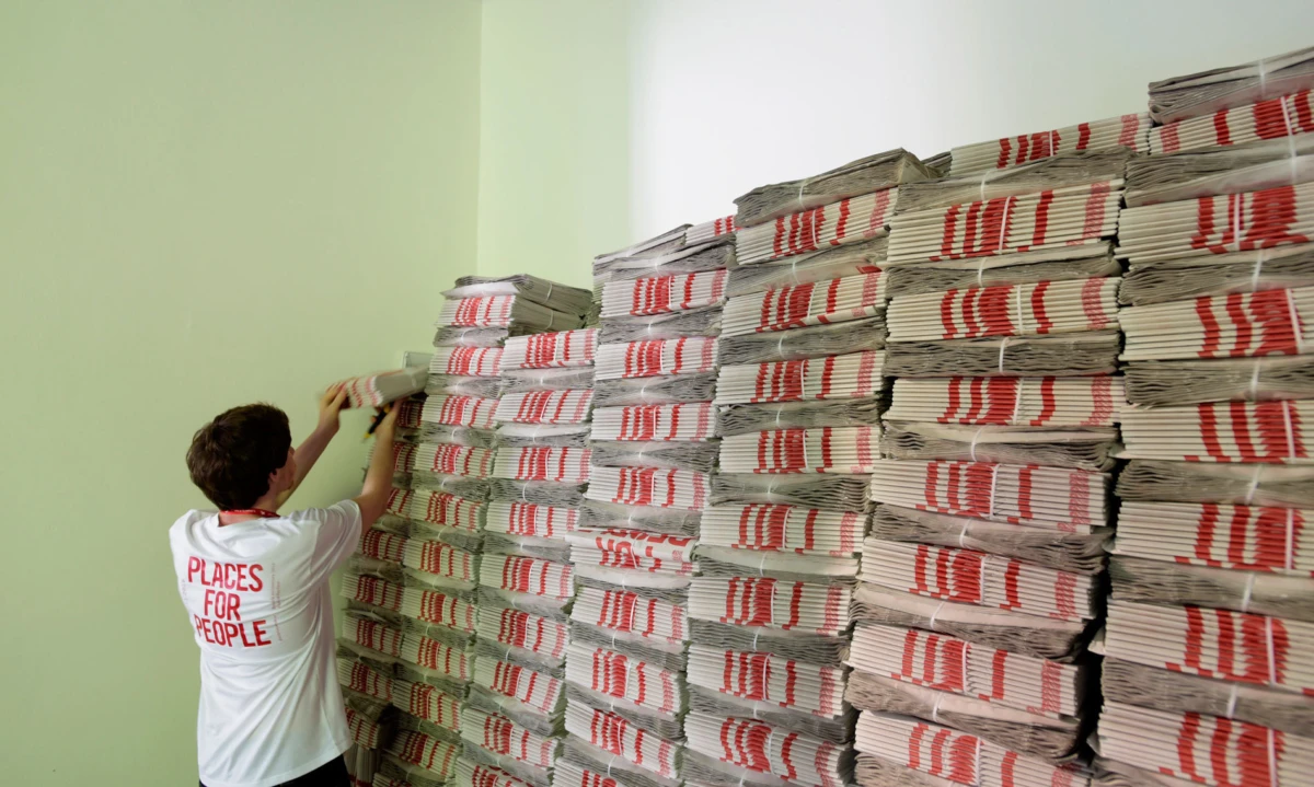 Volunteer arranges stacks of newspapers bound with red-striped wrap against a pale green wall.