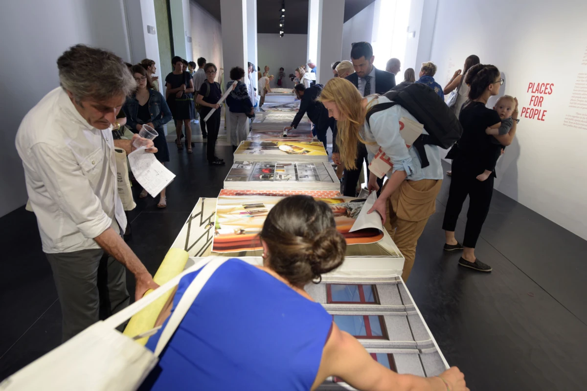 Museum visitors browse large photographic prints laid out on a long table in a contemporary art gallery.
