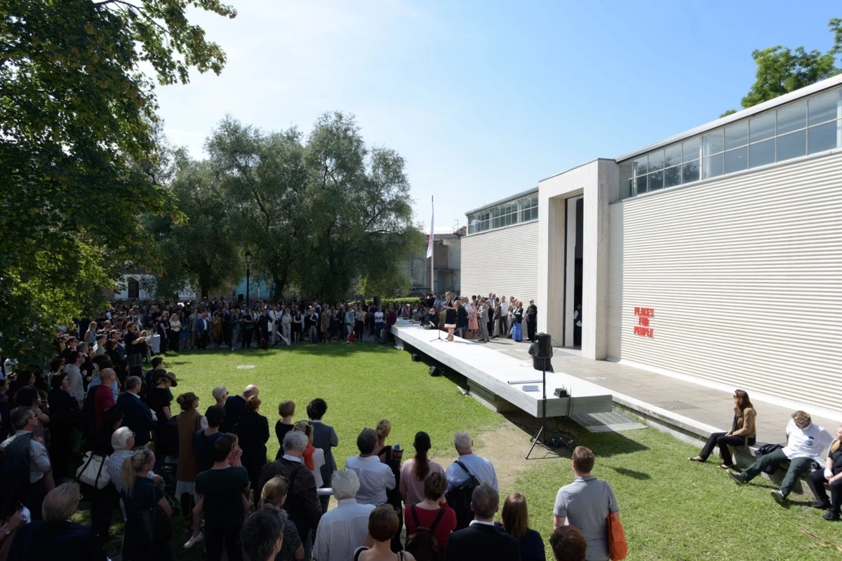 Speaker on a raised platform giving a talk to a large crowd outdoors beside a modern building.