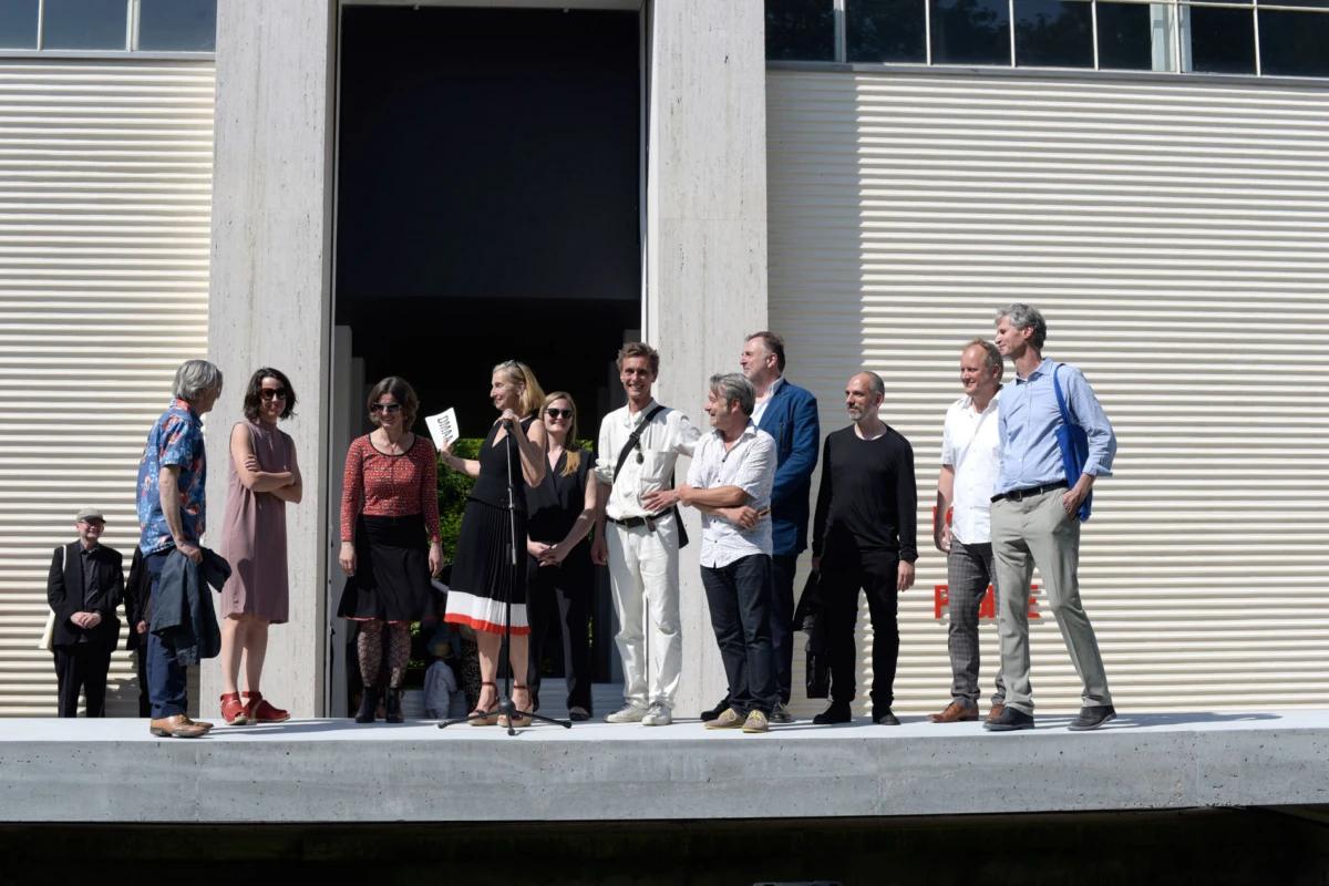 Group of adults on a stage outside a modern building; a woman speaks into a microphone while others stand nearby.