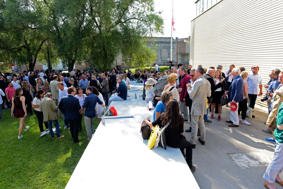 Crowd of attendees mingling at an outdoor reception beside a long white table near a modern building.