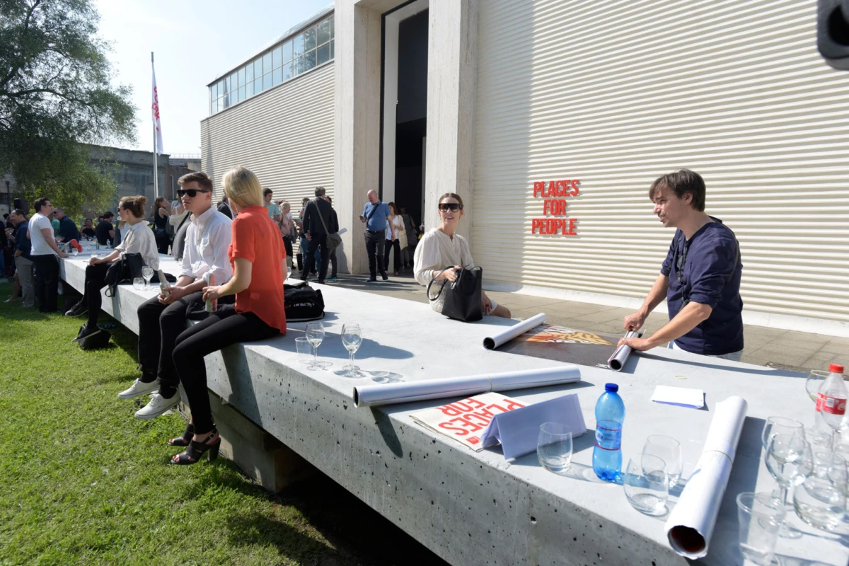 Attendees sit along a long concrete table outdoors as a man unrolls posters beside a modern building.