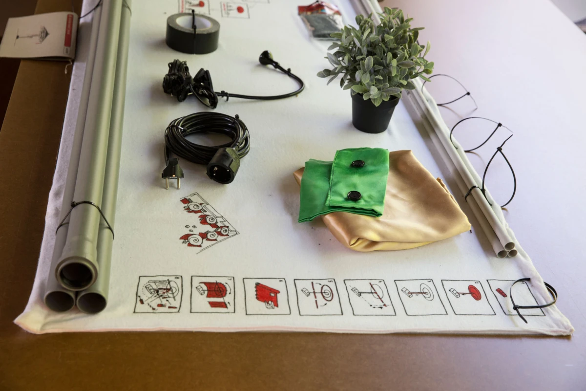 White worktable displaying green-beige fabric swatch with black buttons, a potted plant, coiled power cord, and rolled PVC tubes.