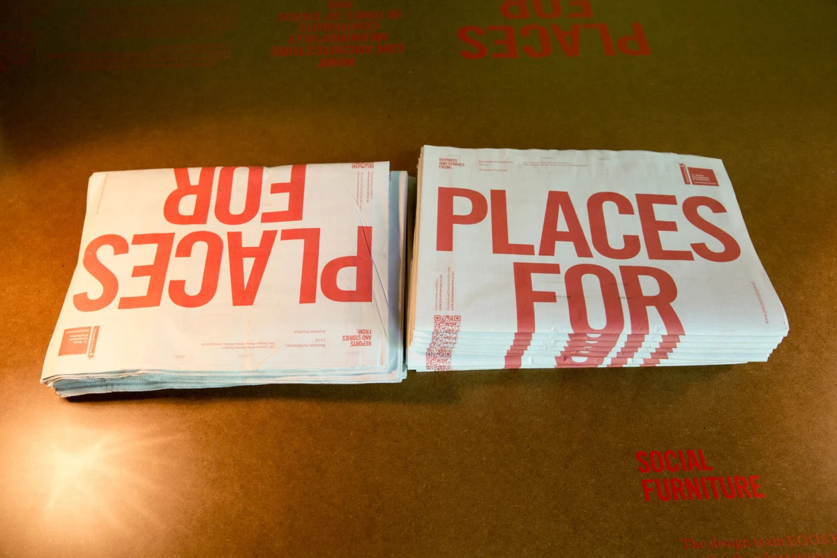 Two stacks of folded newspapers on a brown table, bold red 'PLACES FOR' text visible; 'SOCIAL FURNITURE' also legible.