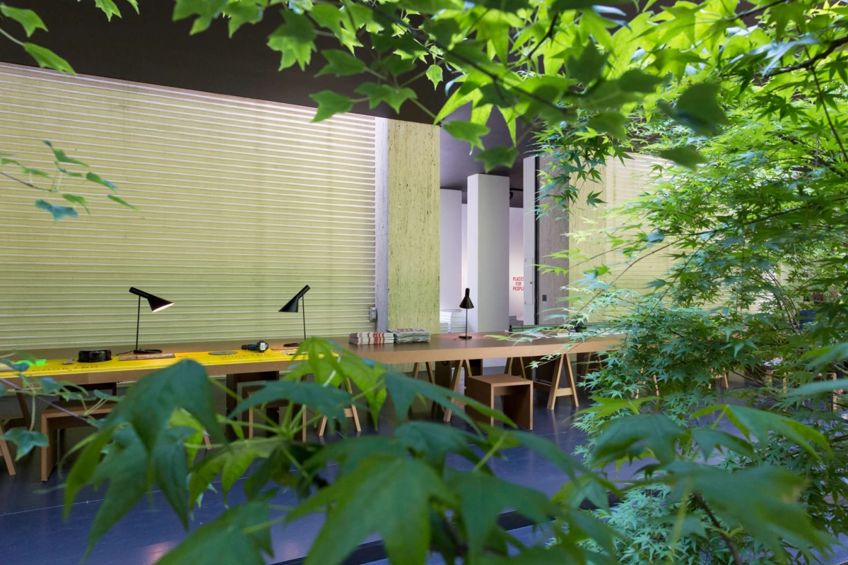 Long wooden worktable with adjustable desk lamps in an indoor biophilic workspace, surrounded by lush green plants.