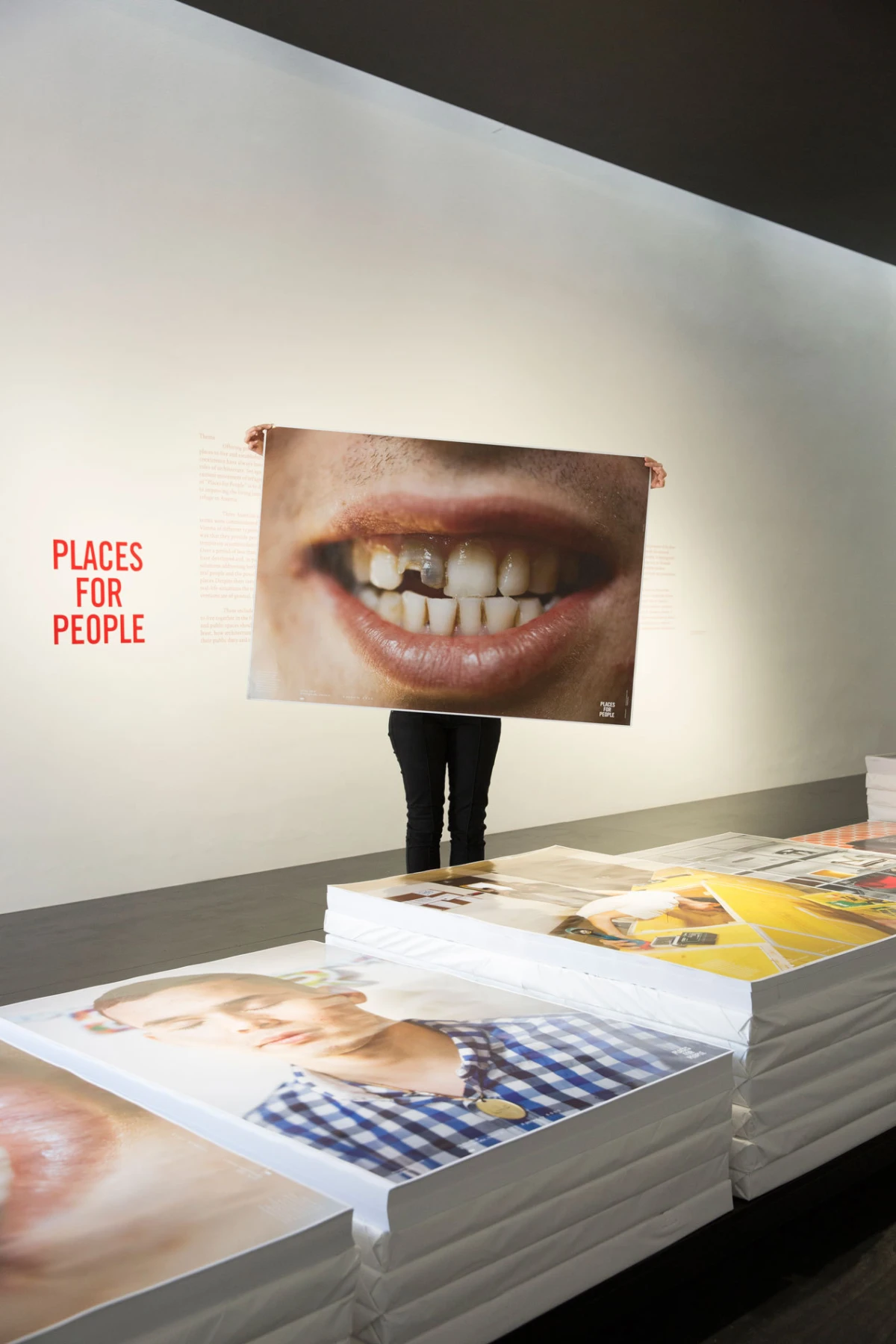Person stands behind a large poster of a close-up smile showing teeth in a gallery; wall text reads PLACES FOR PEOPLE beside stacked portrait prints.