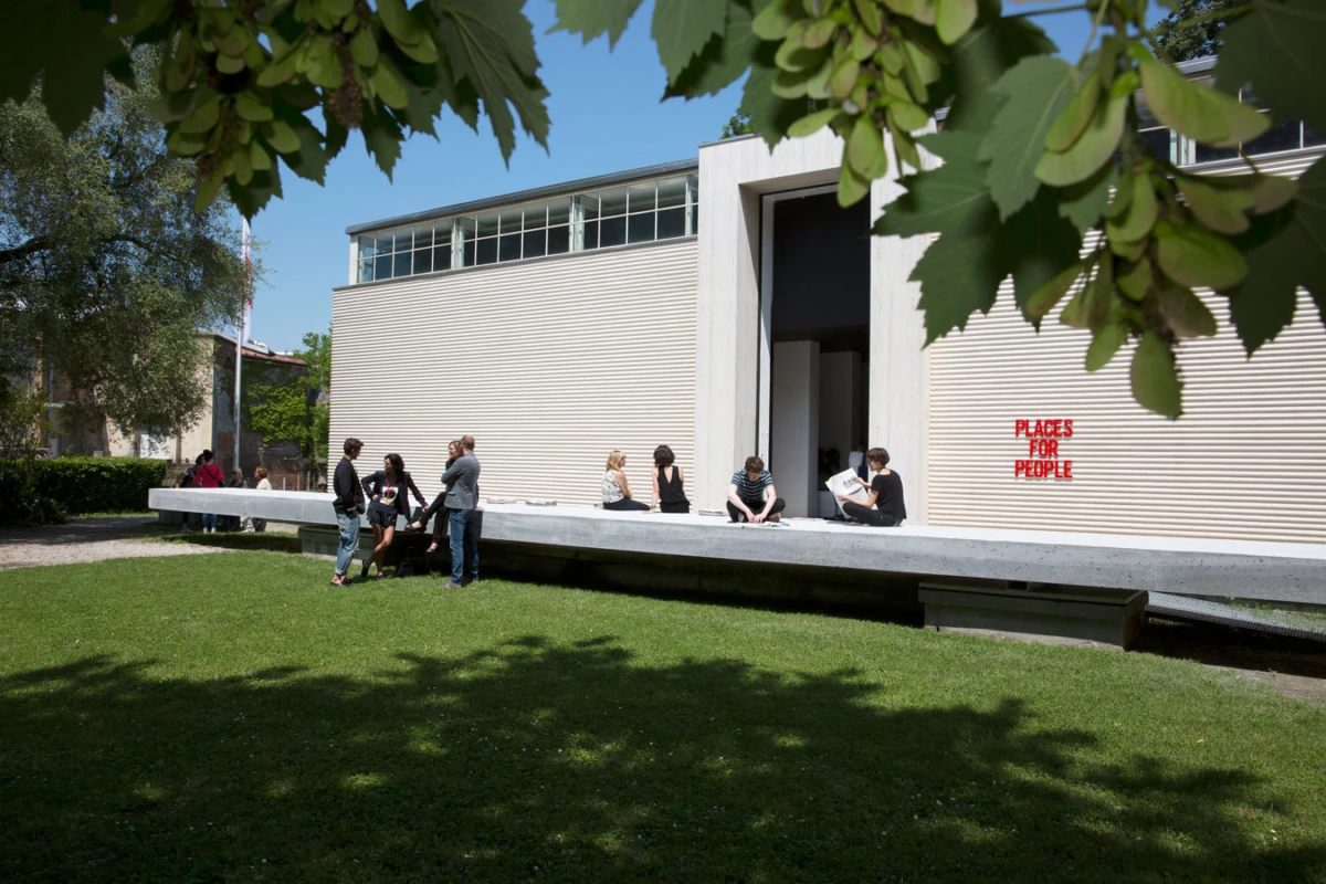 Group of people chatting along a long concrete bench outside a modern beige building; wall sign reads 'PLACES FOR PEOPLE'.