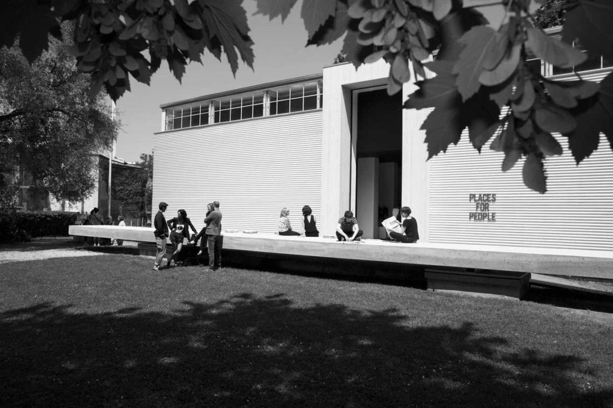 Group of people chat along a long concrete bench outside a modern building with a wide doorway; trees shade the scene.