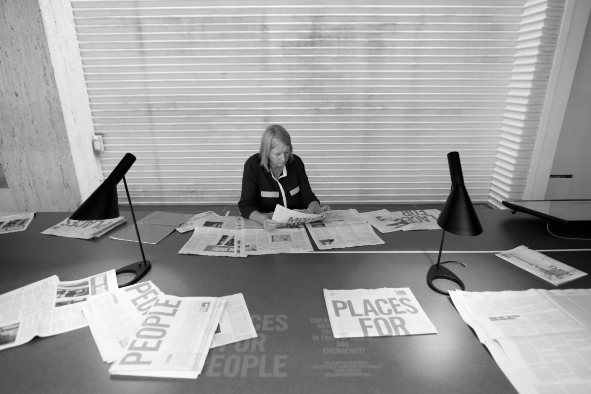 Woman reading a newspaper at a long desk covered with papers, flanked by two desk lamps, with blinds in the background.