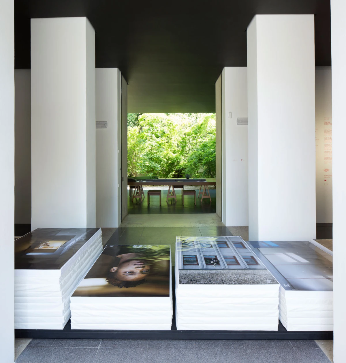 Large photographic prints stacked on the gallery floor, white pillars framing a corridor to a greenery-filled outdoor space.