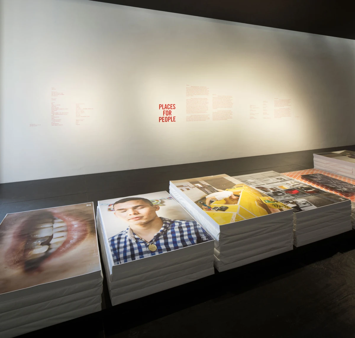 Gallery display of stacked photo prints on white platforms in front of a white wall, central red text PLACES FOR PEOPLE.