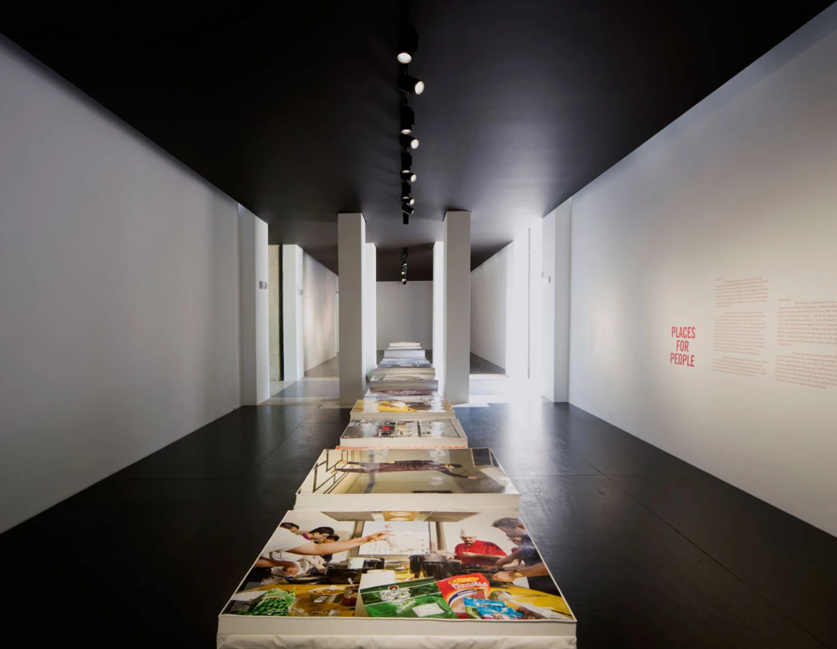Gallery hallway with a central row of photographic panels on white platforms; track lighting and wall text PLACES FOR PEOPLE.