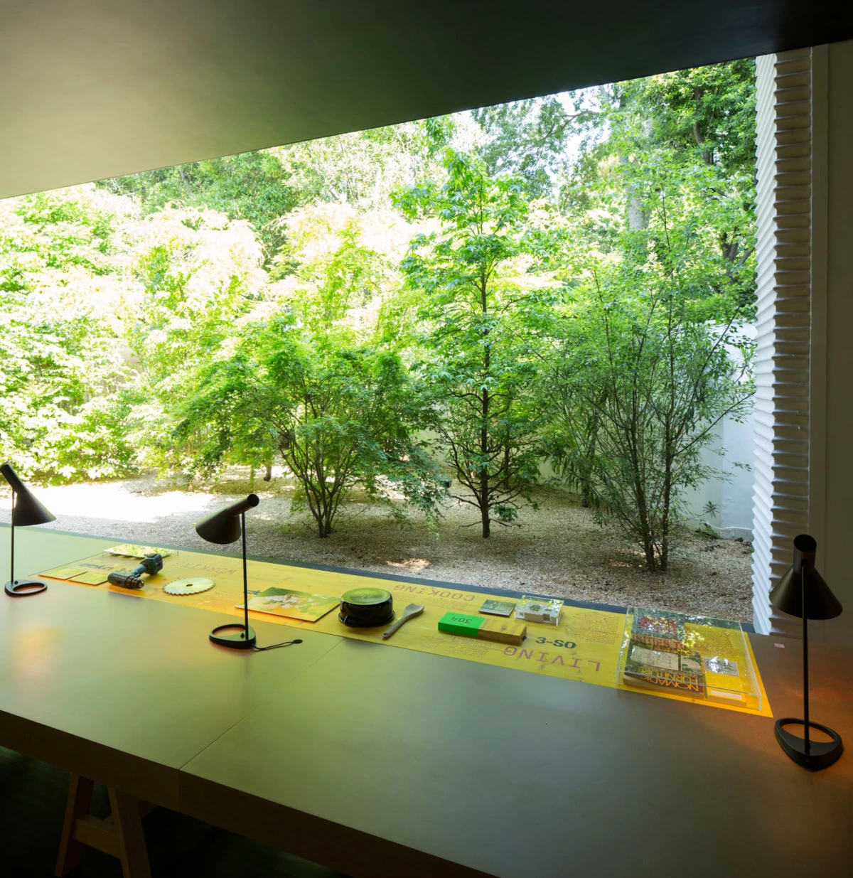 Workbench by a window with two black desk lamps and assorted tools on a yellow mat; lush green garden visible outside.