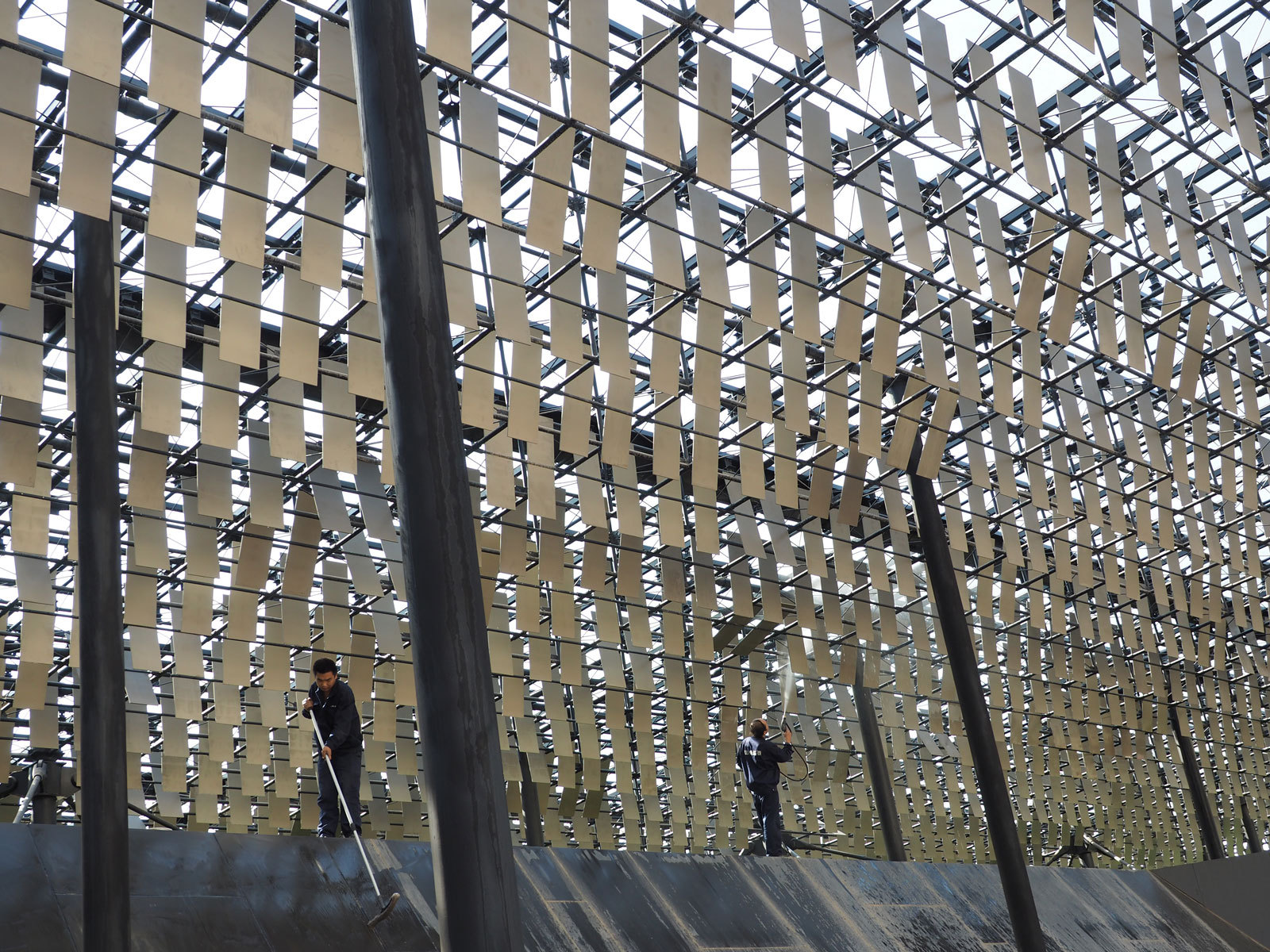 Architectural lattice canopy of beige rectangular panels; two workers cleaning a sloped metal platform beneath.