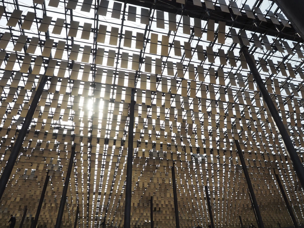 Sunlit outdoor canopy of beige rectangular panels suspended from dark steel columns, forming a grid-like lattice.