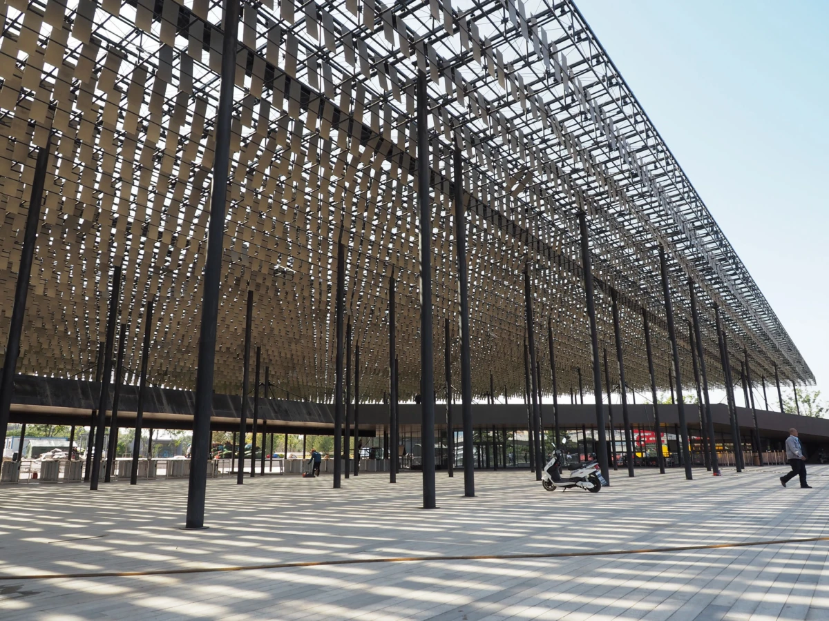 Outdoor plaza under a lattice metal canopy supported by tall black columns; a scooter parked and a pedestrian walking nearby.