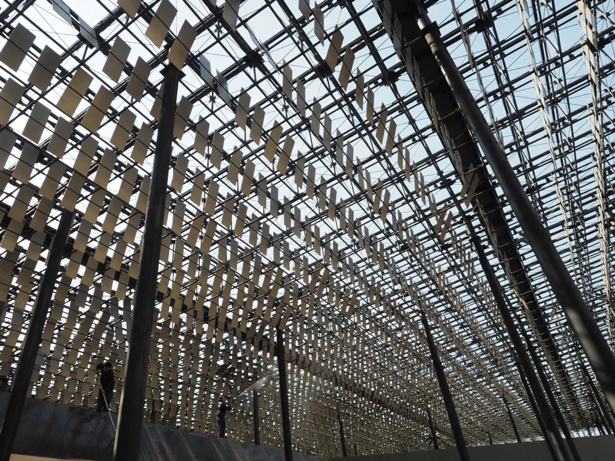 Glass-roofed atrium with hundreds of beige rectangular panels suspended from a metal grid; people walk along a lower walkway.