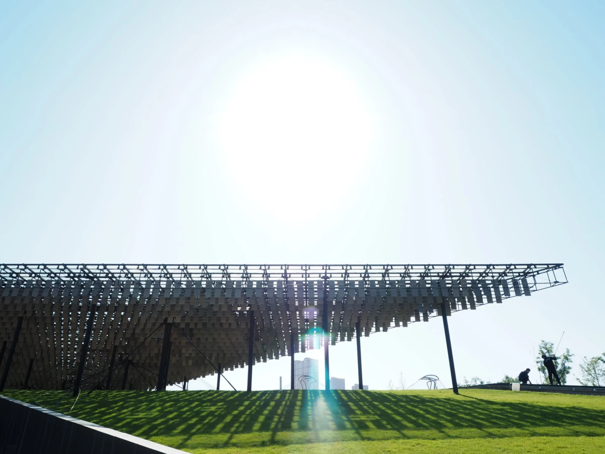 Steel lattice canopy over a grassy hill; bright sun overhead, with two people standing near the edge.