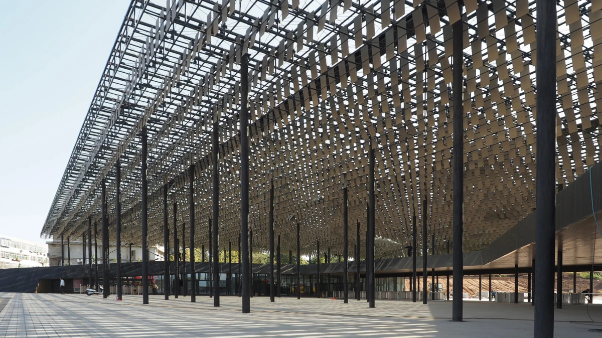 Open-air pavilion with a lattice metal roof supported by tall black columns over a spacious tiled plaza.