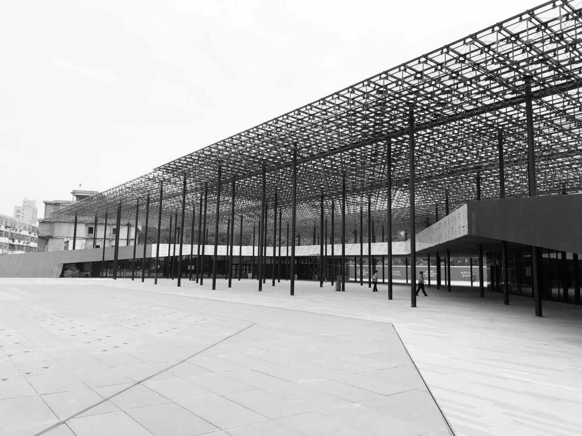 Open plaza with a tall metal lattice canopy supported by vertical columns; several pedestrians walk beneath a modern building.