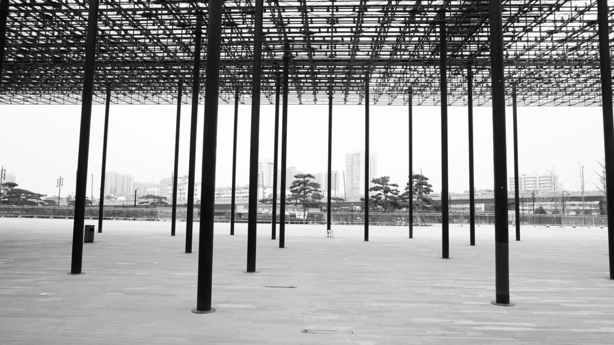 Rows of tall steel columns support a lattice roof over an empty plaza, with an urban skyline in the distance.