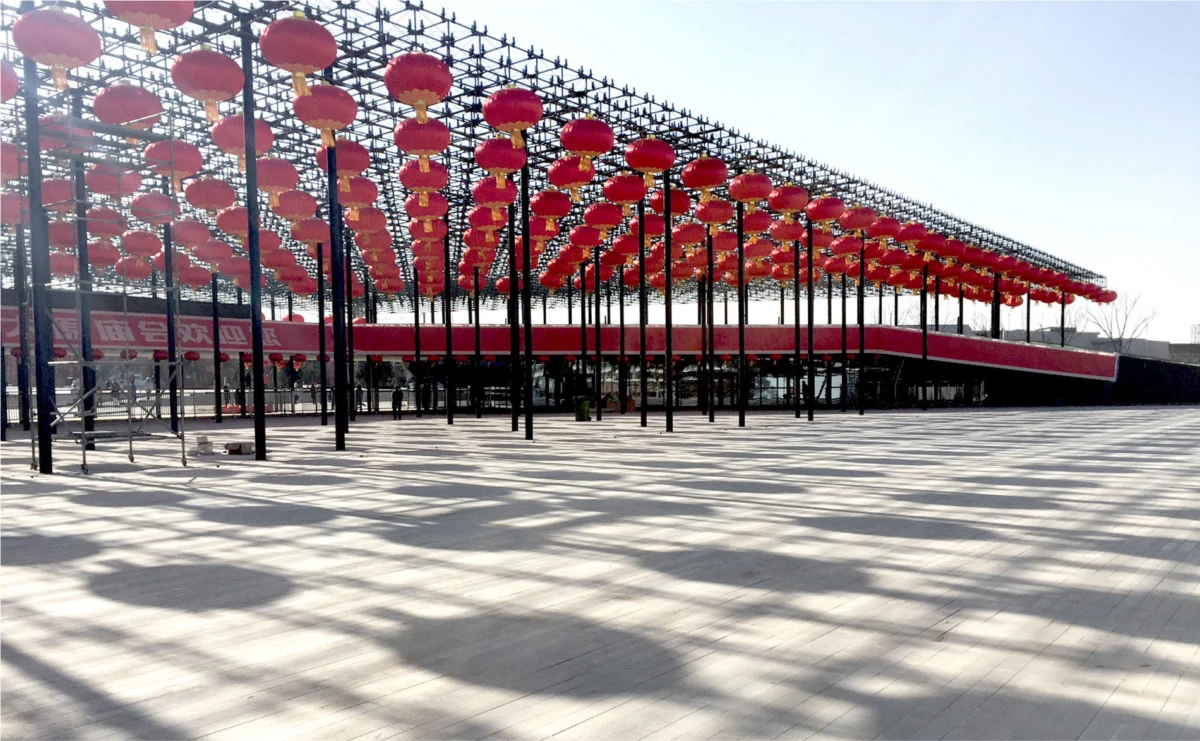 Rows of red Chinese lanterns hang above a covered outdoor plaza with black supporting poles, creating striped shadows on the concrete square.
