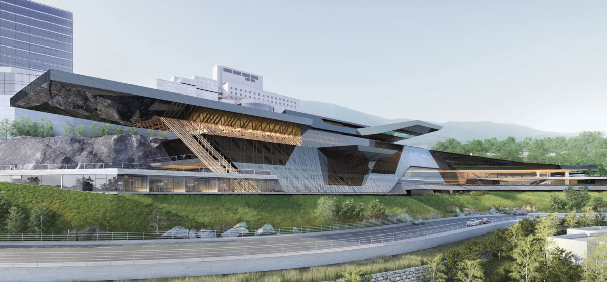 Futuristic angular building perched on a grassy hillside beside a highway, with cars in the foreground and distant mountains.