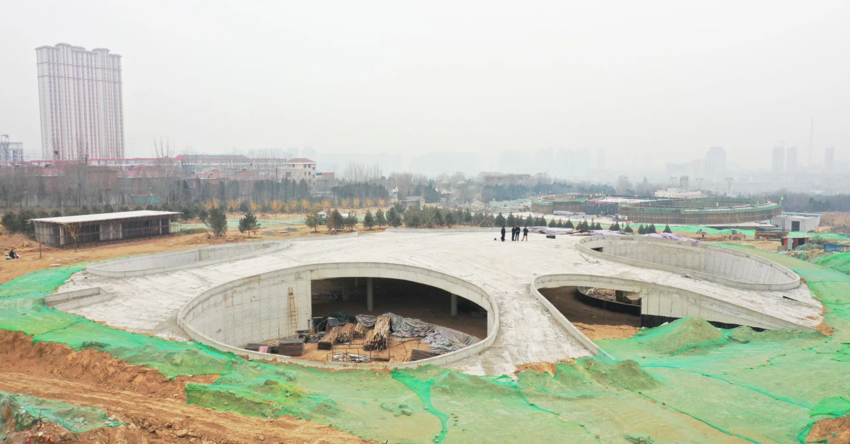 Circular concrete structure under construction at an urban site; workers on the rim, green tarps and dirt around, hazy city skyline in distance