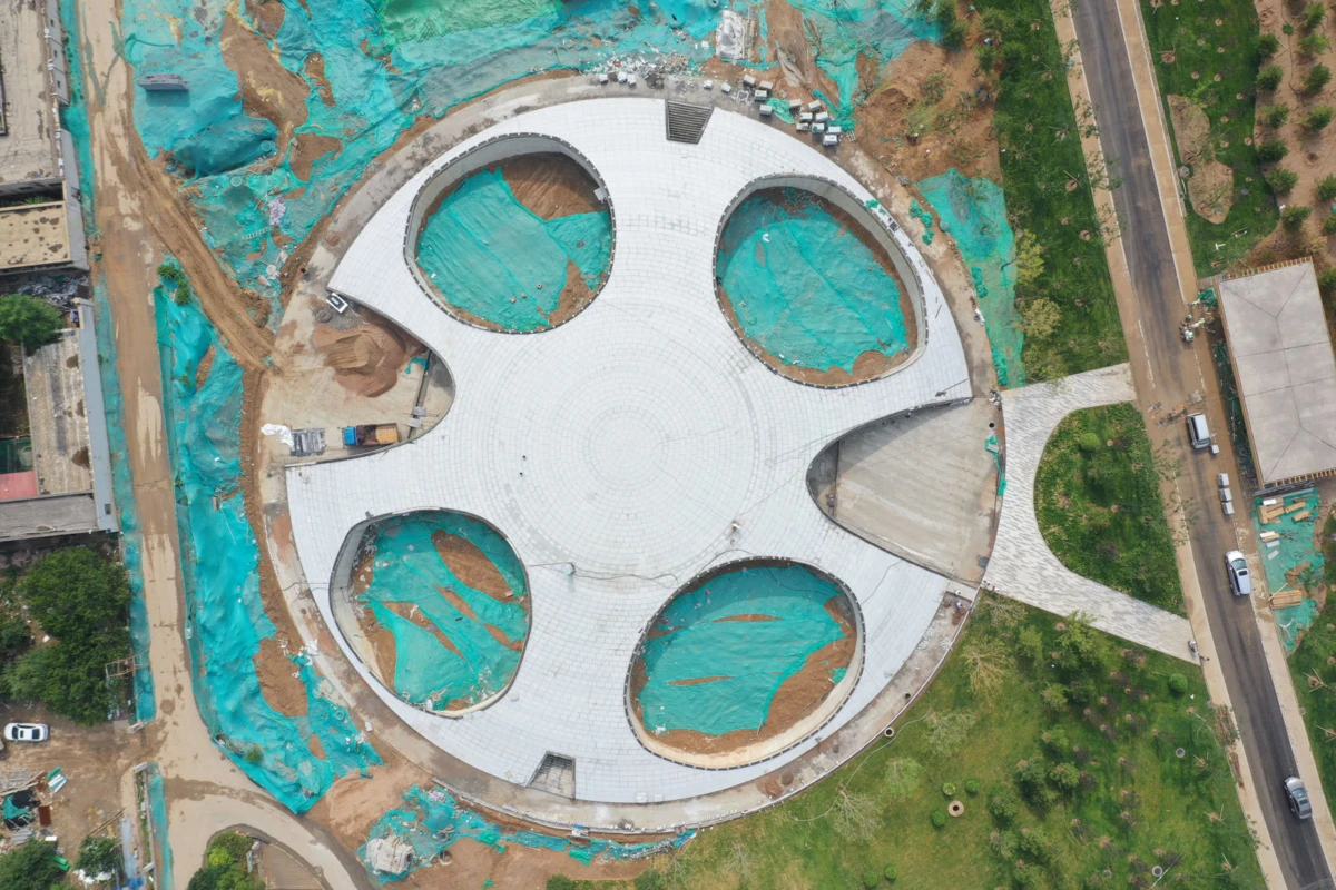 Aerial view of a circular plaza under construction with four circular cutouts, white paving, turquoise tarps, and surrounding roads and greenery.
