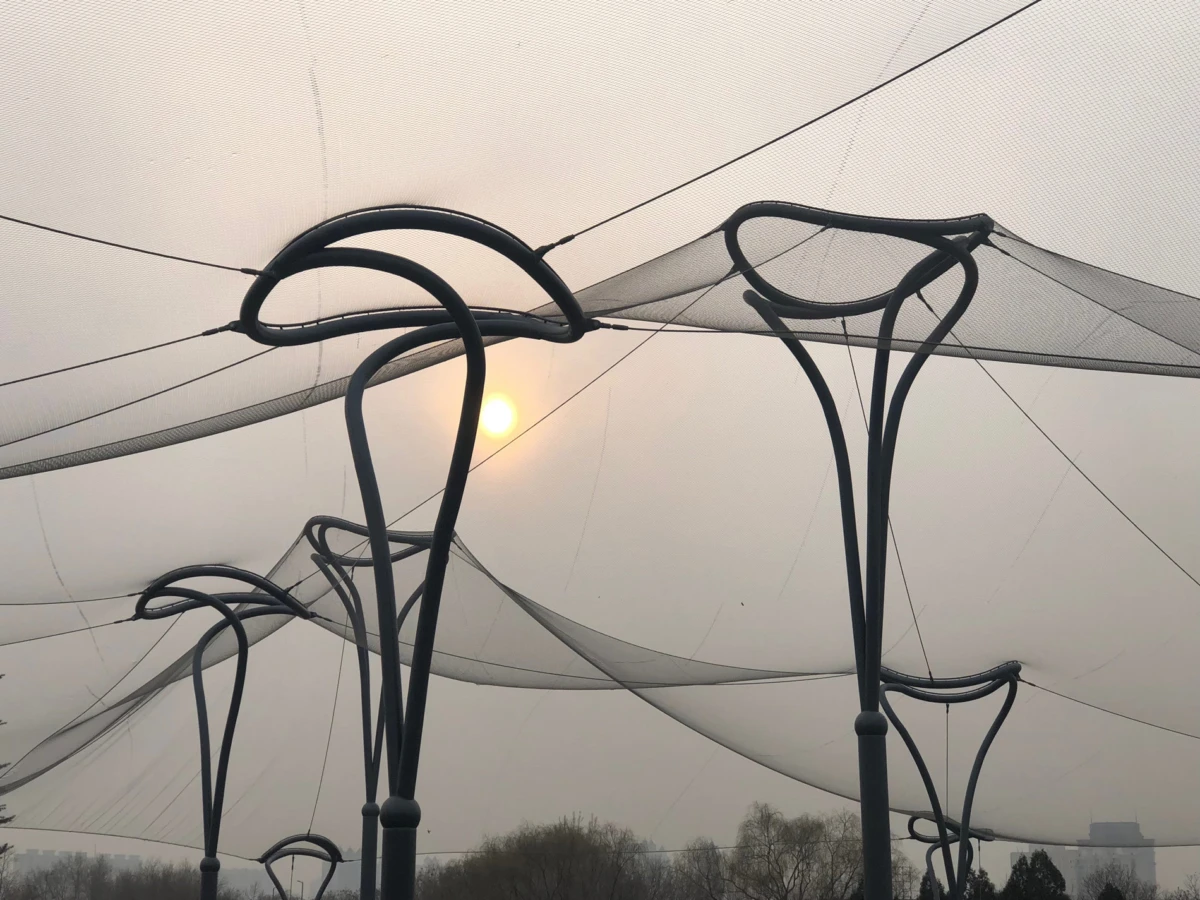 Abstract black metal arches form looping shapes as netting is draped overhead in a hazy outdoor park at sunset.