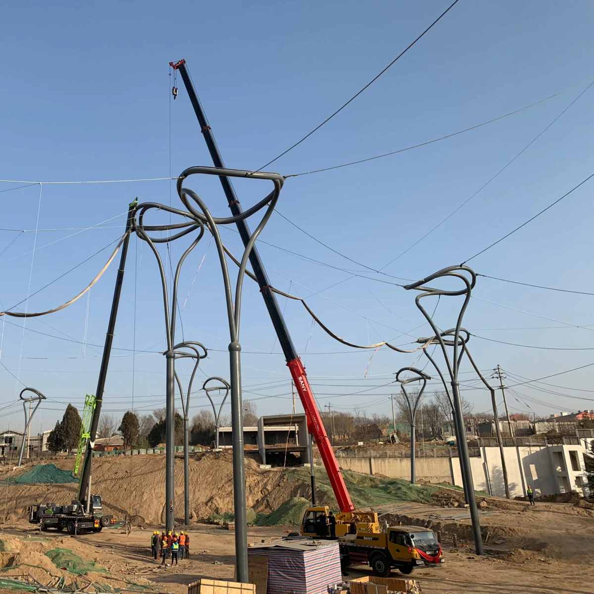 Crane lifts curved steel framework for overhead lines at a construction site; tall looped poles with workers on dirt ground under a blue sky.