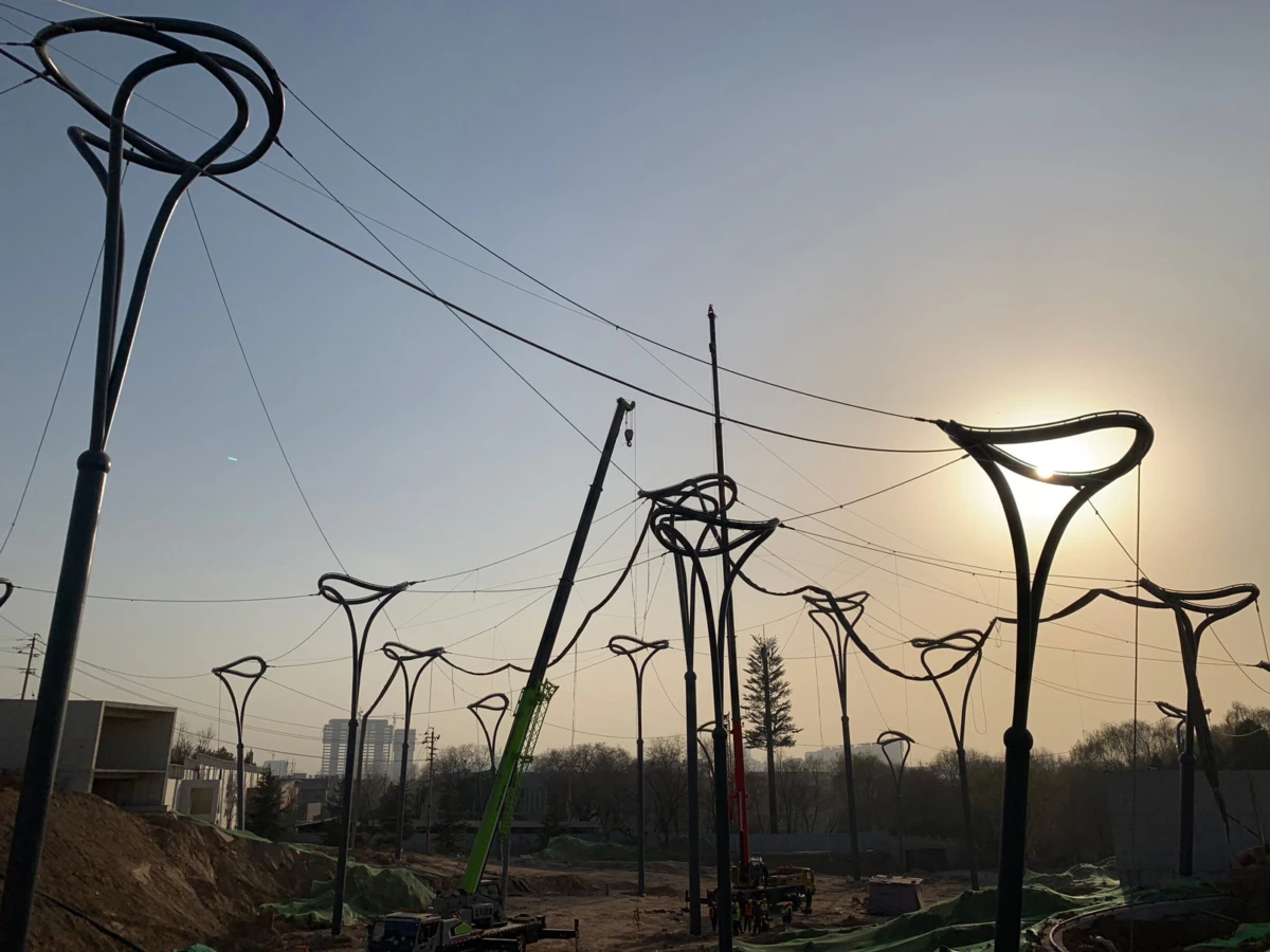 Tall curved streetlight poles with looped tops under construction at a site, with cranes and dirt piles, sunset behind.