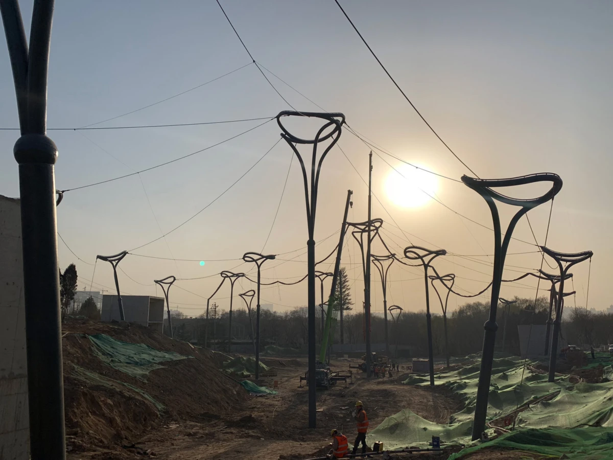 Construction site with tall curved streetlight poles, workers in orange safety vests, dirt mounds and green tarps at sunset.