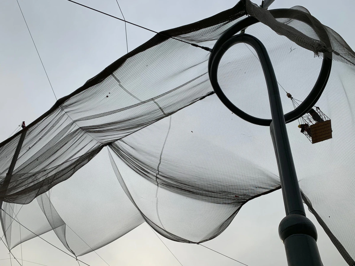 Wind-swept black mesh netting suspended between cables above an urban area, with a worker in a suspended basket repairing it near a curved metal post.