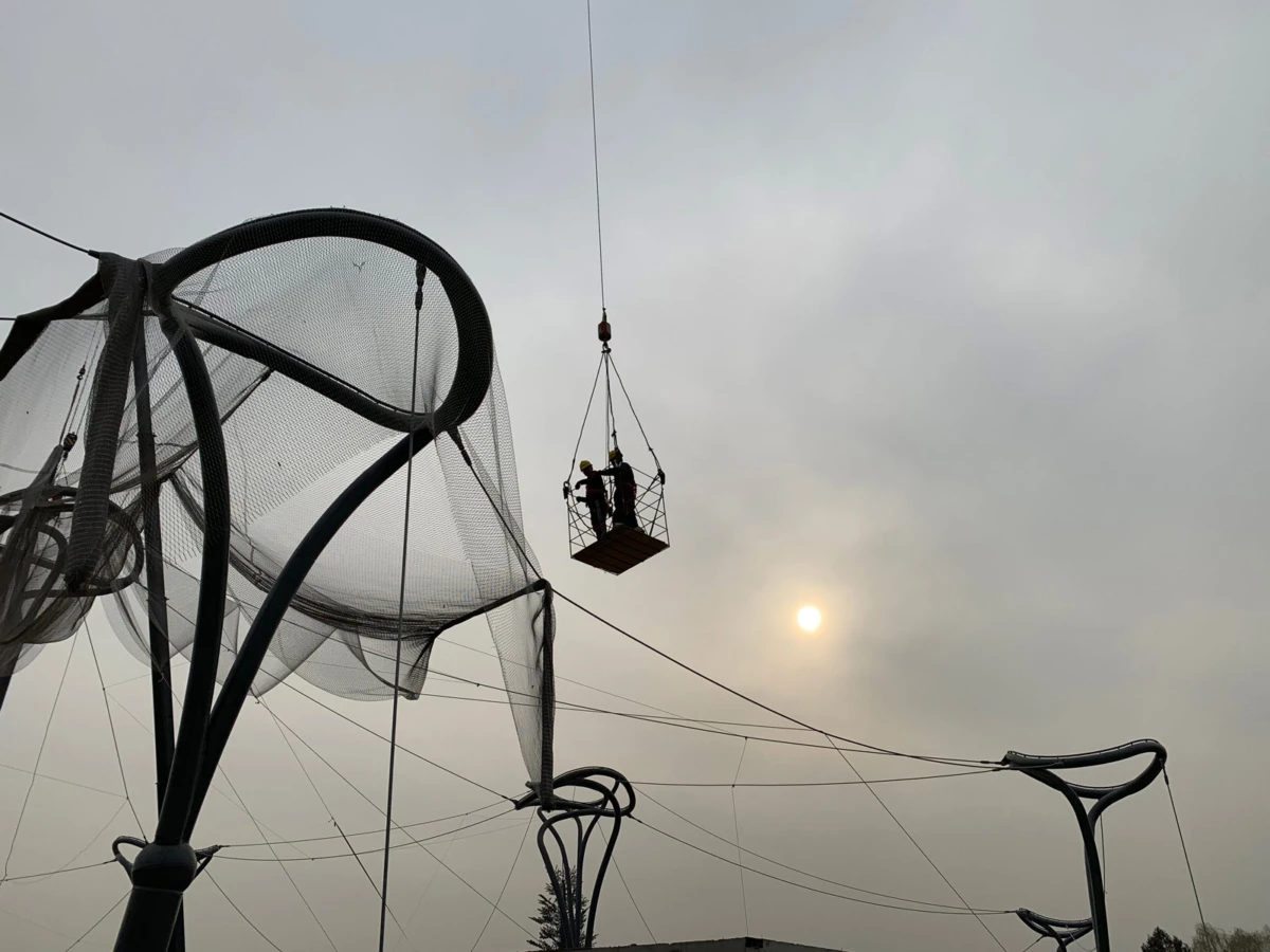 Two workers on a suspended work basket hoisted by a crane, tending a large circular netted sculpture outdoors under a hazy sky.