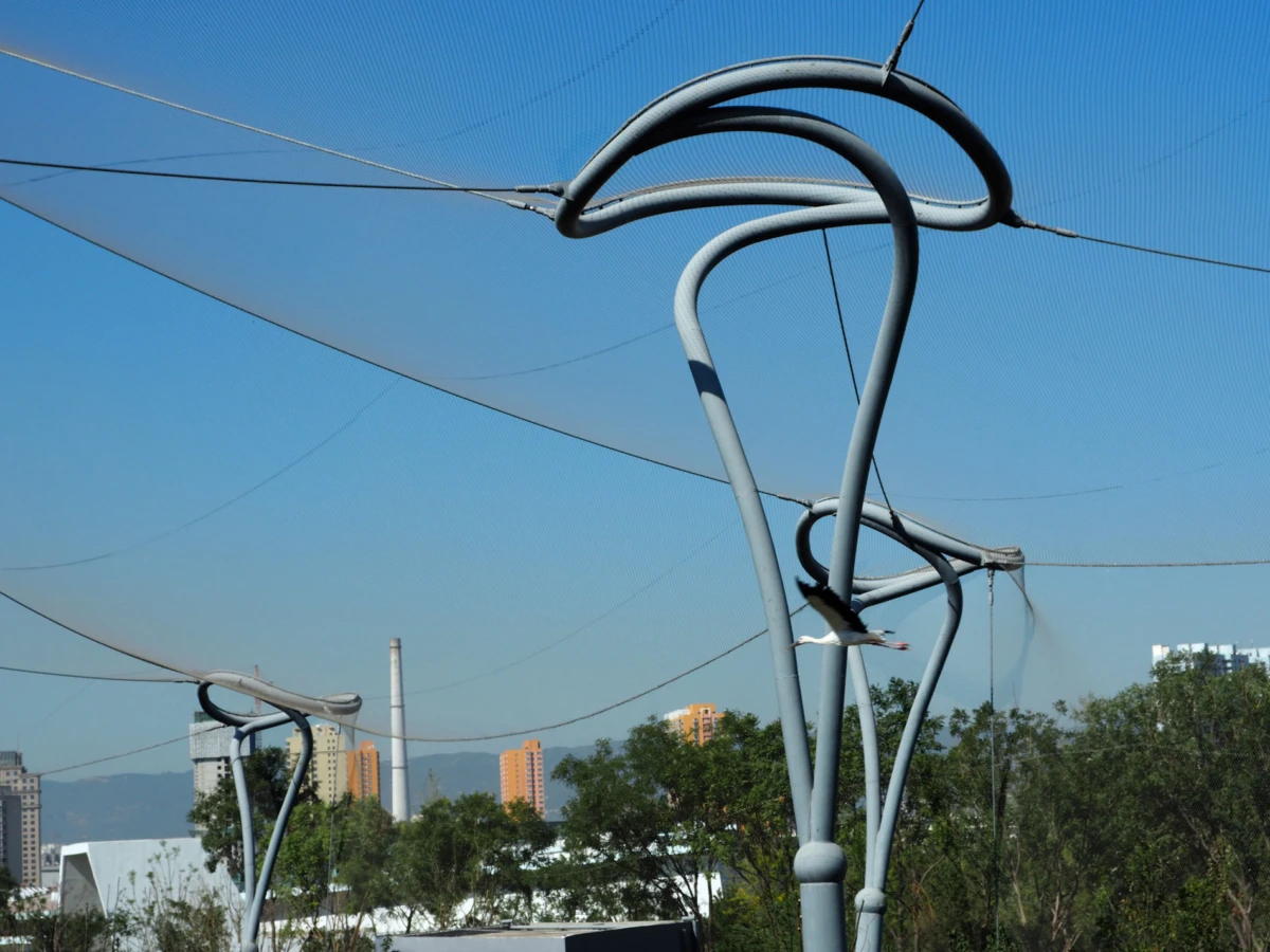 Tubular steel arches support overhead netting over an urban backdrop with trees and high-rise buildings.