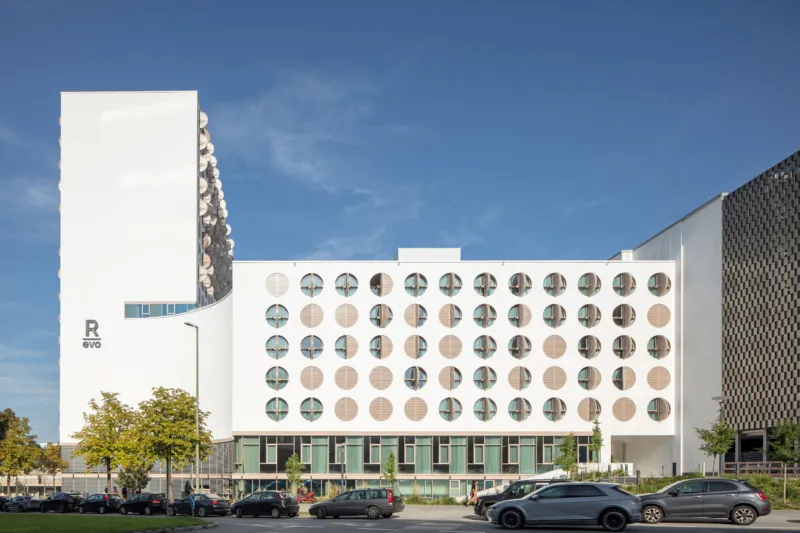 White modern office building with multiple circular windows, cars parked along the street in front, blue sky and trees.