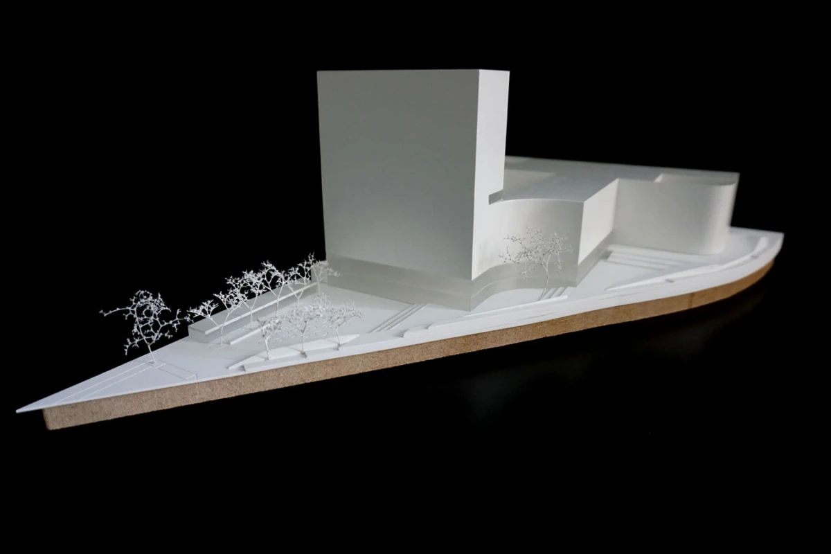 White architectural scale model featuring a tall rectangular block and curved volumes on a base, with small trees along a curved street.