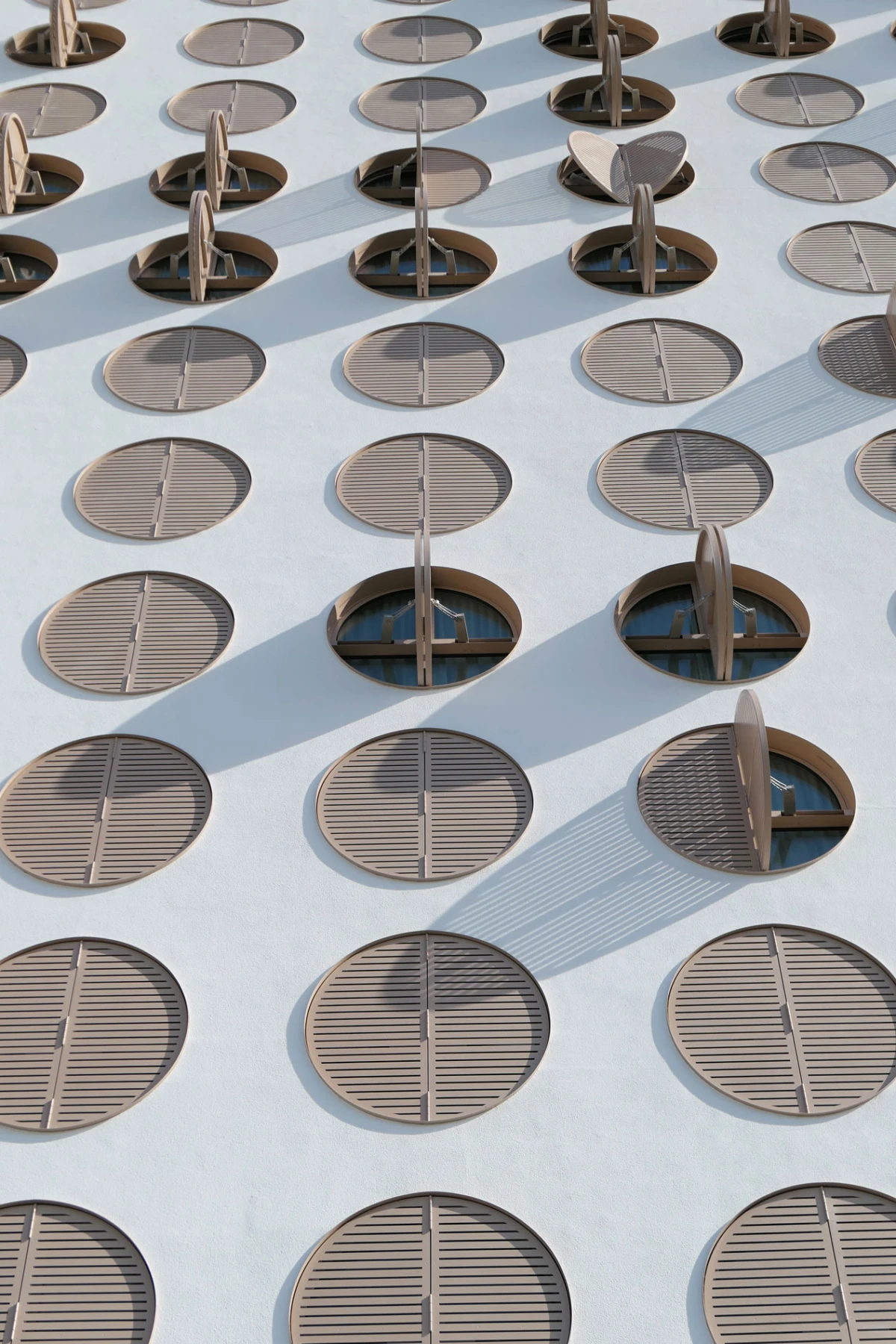 Exterior wall featuring a grid of brown circular louvers, some partially open, casting long diagonal shadows.
