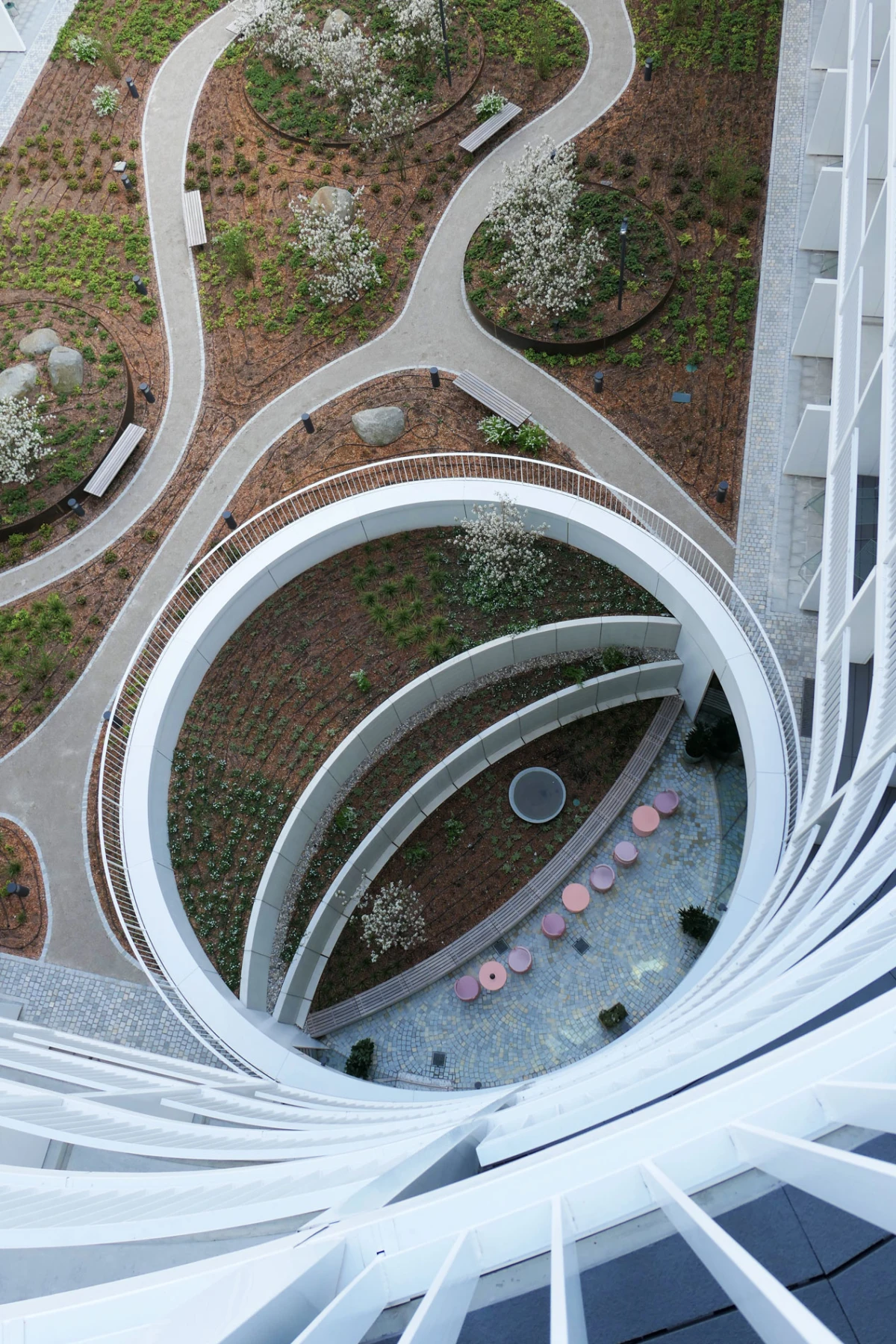 Aerial view of a spiraling white atrium with concentric circular ramps around a garden courtyard and pink circular seating along a tiled plaza.